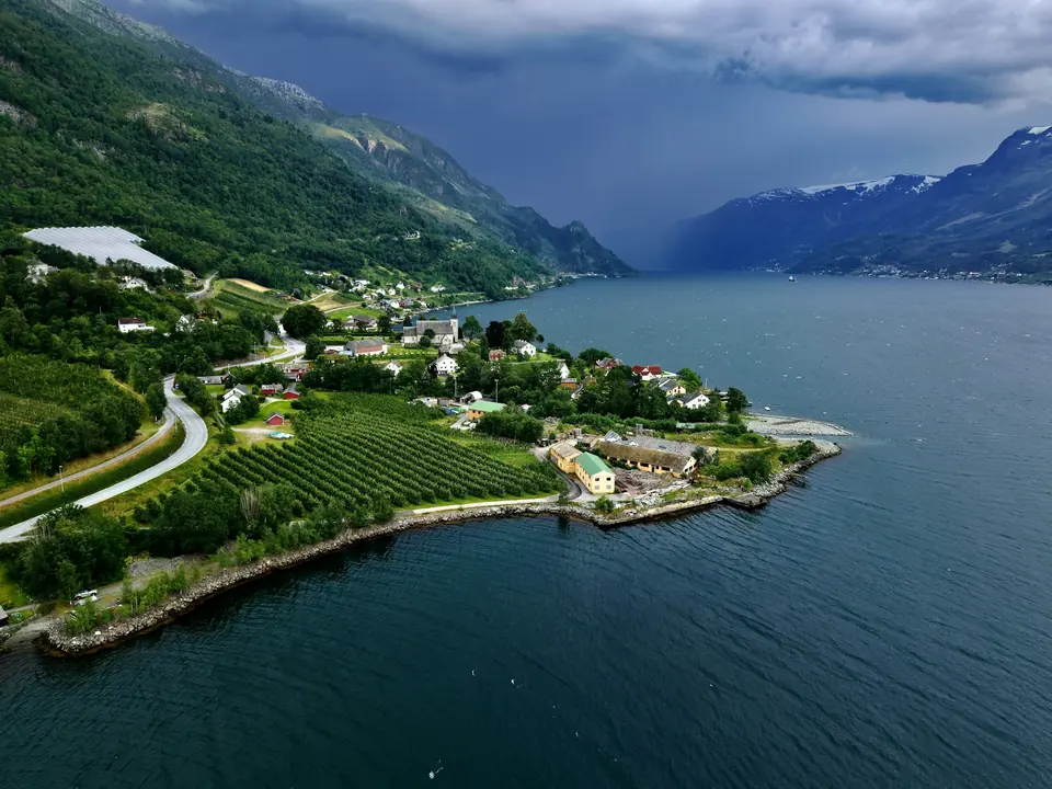 Fjordbygd med frukthager, gårder og en svingete vei sett ovenfra under mørke skyer